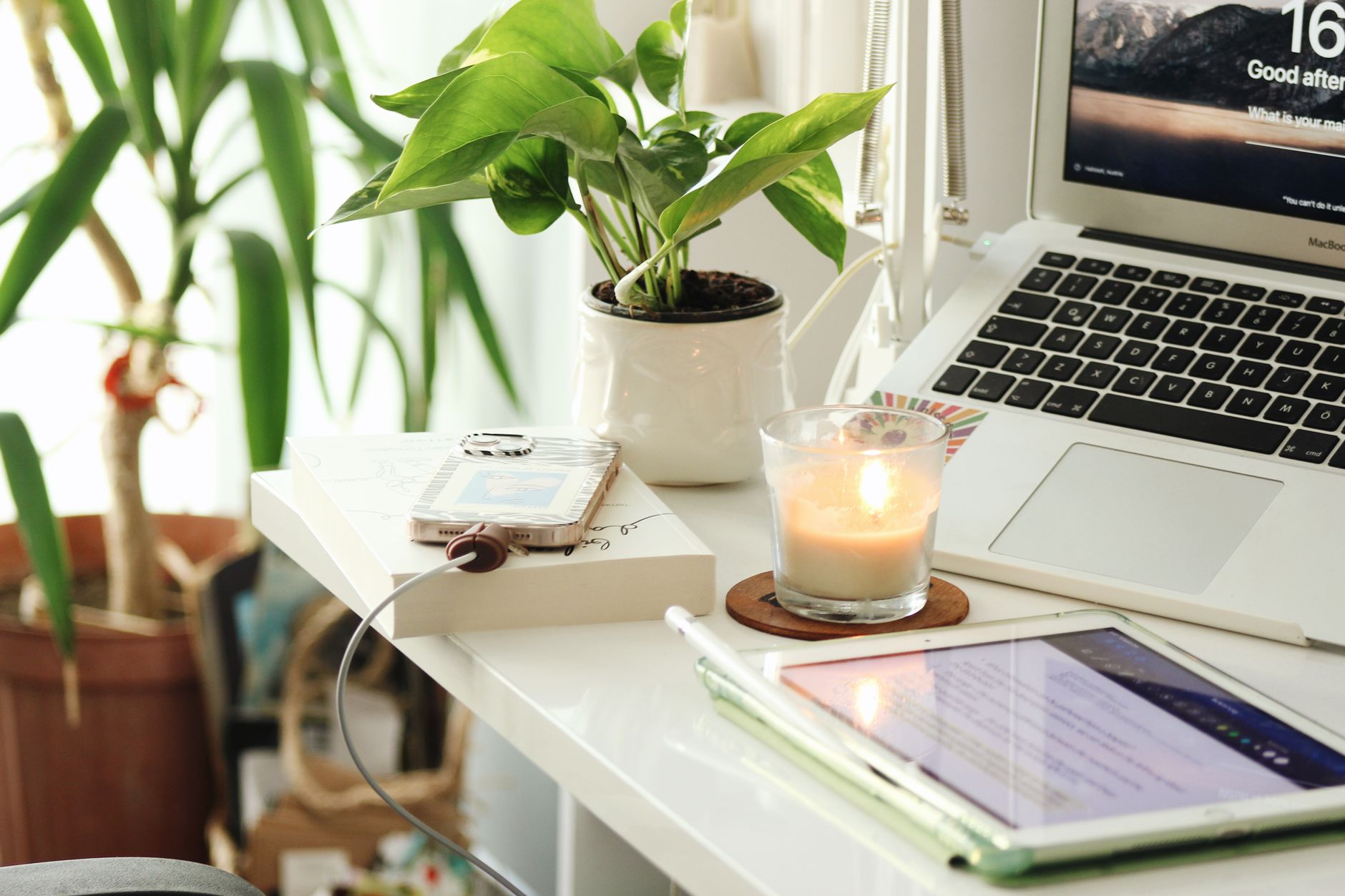 A bright home desk with an open laptop, a tablet, a phone, a lit candle and a potted plant.