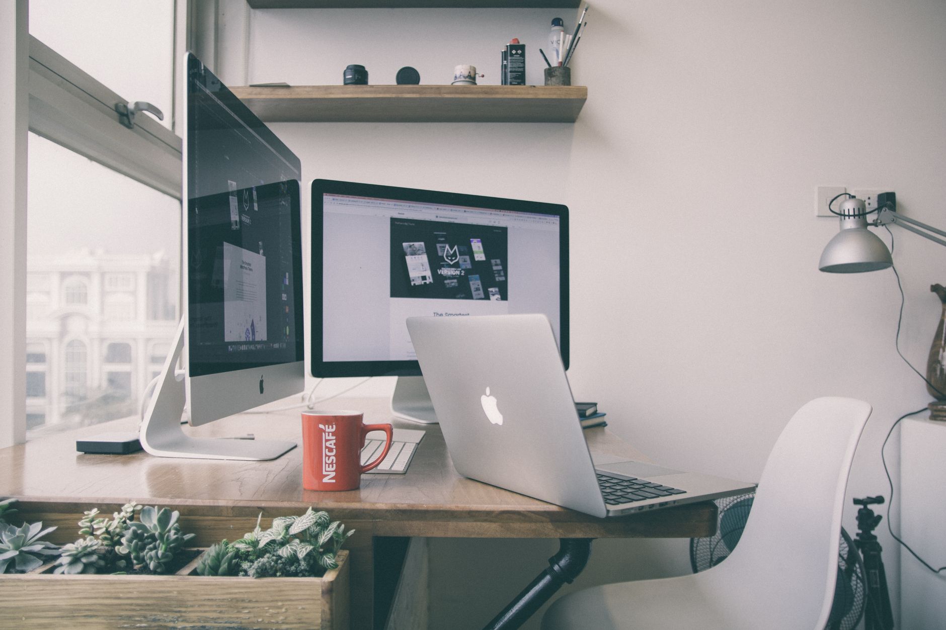 A home office desk with two monitors, a laptop, a red mug and a white chair beside a window.