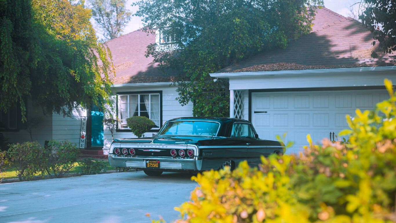 Voiture ancienne noire garée dans l’allée d’une maison de quartier résidentiel, entourée de feuillage