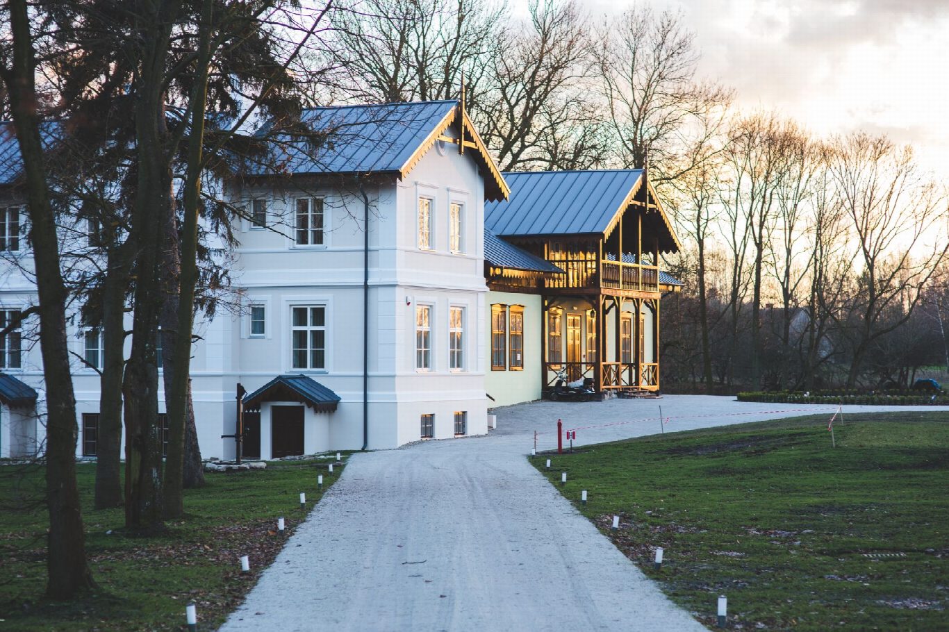 Grande maison blanche à toit sombre au bout d’une allée gravillonnée, au coucher du soleil, entourée d’arbres