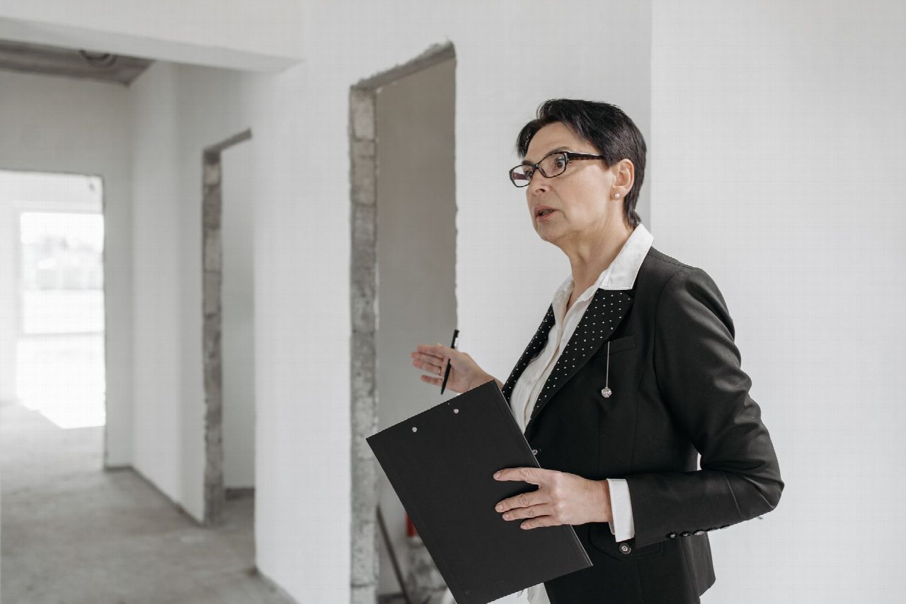 Une femme en tailleur noir tient un bloc-notes et observe un local en rénovation aux murs encore bruts.